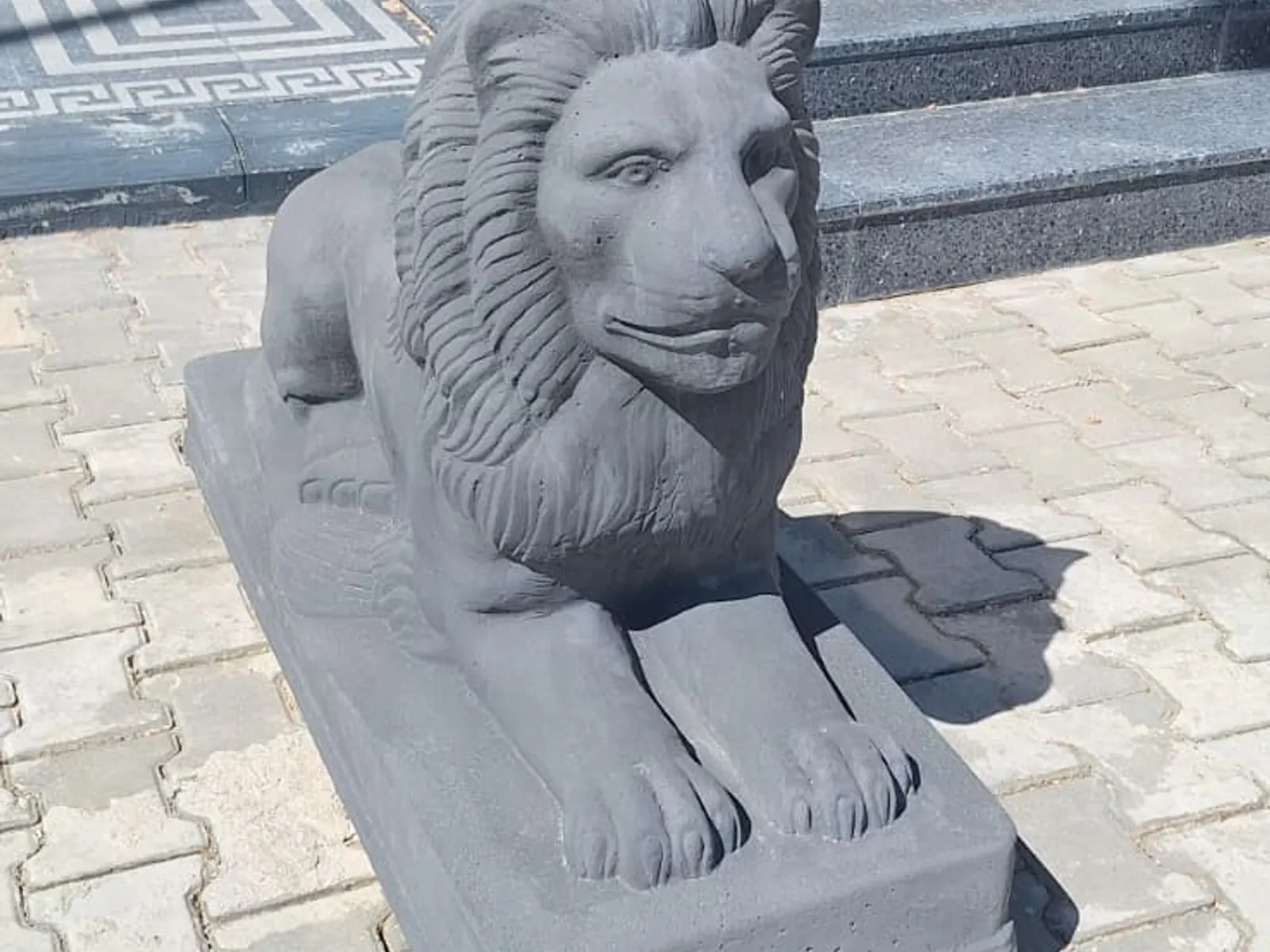 Beton aslan figürünün açılı çekimi, arkada tesis girişi ve granit basamaklar
