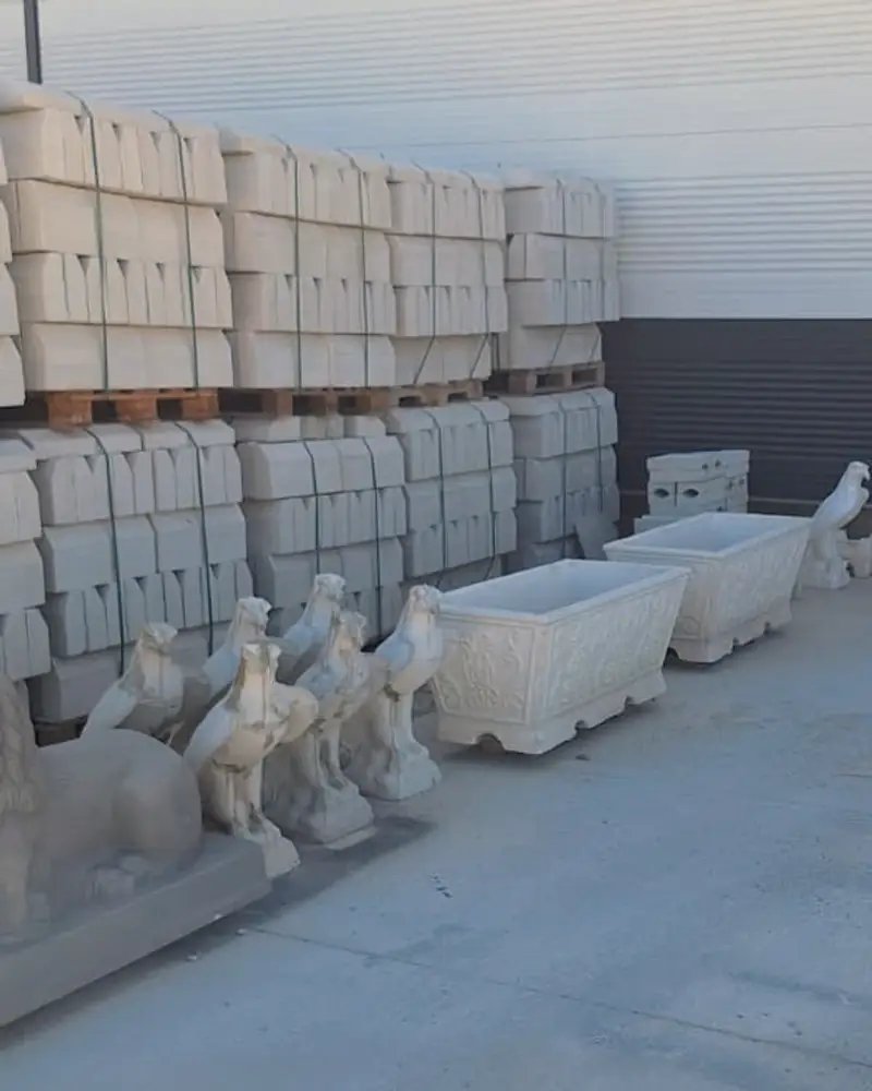 Tesiste paletlenmiş beton bordür stoğu, önünde beton kuş ve aslan figürleri ile kabartmalı çiçeklikler